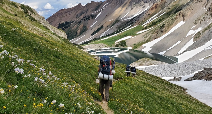 Students carrying backpacks hike along a trail in a grassy open valley between snow-covered mountains